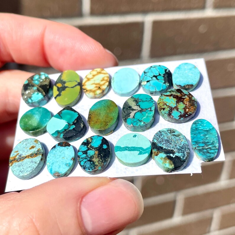 May include: A collection of turquoise gemstones in various shapes and sizes, including oval and round. The stones display a range of colors, from light blue to deep teal, with brown and black matrix patterns. The gemstones are displayed on a white surface.