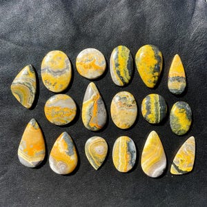 May include: A collection of 17 yellow, black and grey bumblebee jasper cabochons. The cabochons are various shapes and sizes, with some being teardrop shaped and others being oval shaped.