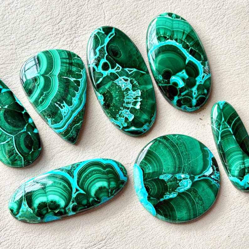 Pu&ograve; includere: Una collezione di sei pietre preziose di malachite lucidata in varie forme, tra cui ovali, rotonde e a goccia. Ogni pietra mostra bande concentriche e intricati motivi a vortice nei toni del verde bosco e del verde smeraldo.