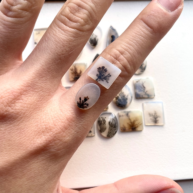 May include: A close-up of a hand holding two white agate cabochons with black dendritic patterns. The cabochons are set in a square and oval shape.