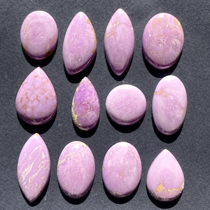 May include: A collection of twelve polished gemstones in various shapes, including oval, teardrop, and elongated forms. The stones are a soft lavender color with subtle golden veining, set against a dark background.