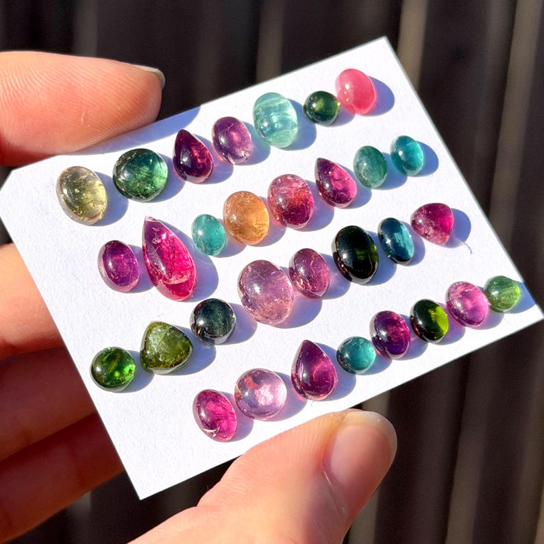 May include: A collection of small, polished, multi-colored tourmaline gemstones arranged on a white card. The stones feature various shapes including oval, round, and teardrop, displaying vibrant shades of pink, green, blue, and yellow.