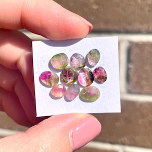 May include: A collection of ten small, oval-shaped, multi-coloured gemstones. The gemstones are a mix of pink, green, and black.