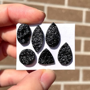 May include: Six black tektite gemstones in different shapes: oval, leaf, teardrop, round, and triangular. The tektites have a textured, glassy look and are shown on a white card. A brick wall forms the background.