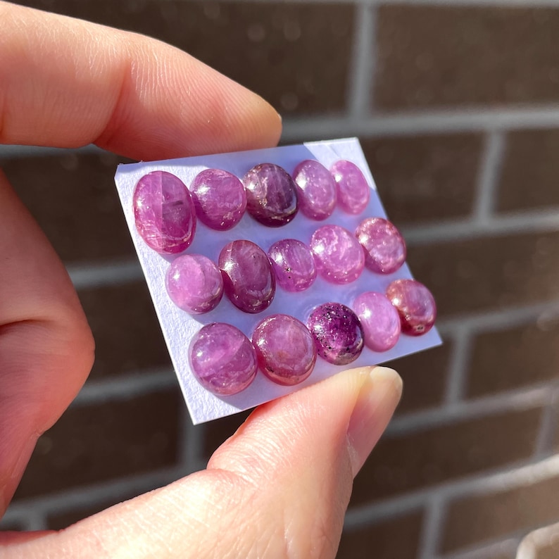 Pode incluir: Uma cole&ccedil;&atilde;o de pedras preciosas ovais polidas em tons variados de rosa e roxo. As pedras est&atilde;o dispostas em um cart&atilde;o branco, segurado por uma m&atilde;o. As pedras t&ecirc;m uma superf&iacute;cie lisa e reflexiva, provavelmente para joias ou artesanato.