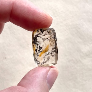 May include: A clear, rectangular gemstone with black, yellow, and brown inclusions, like a tiny landscape. The polished stone is held between fingers, revealing its unique patterns. The gemstone's dimensions are not visible.
