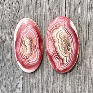 Op de afbeelding: Twee ovale roze en witte gebande agaat cabochons. De cabochons hebben een draaiende patroon van roze, wit en bruin.