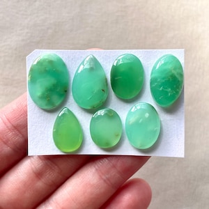 May include: Seven green gemstone cabochons in various oval and teardrop shapes, displayed on a white card.