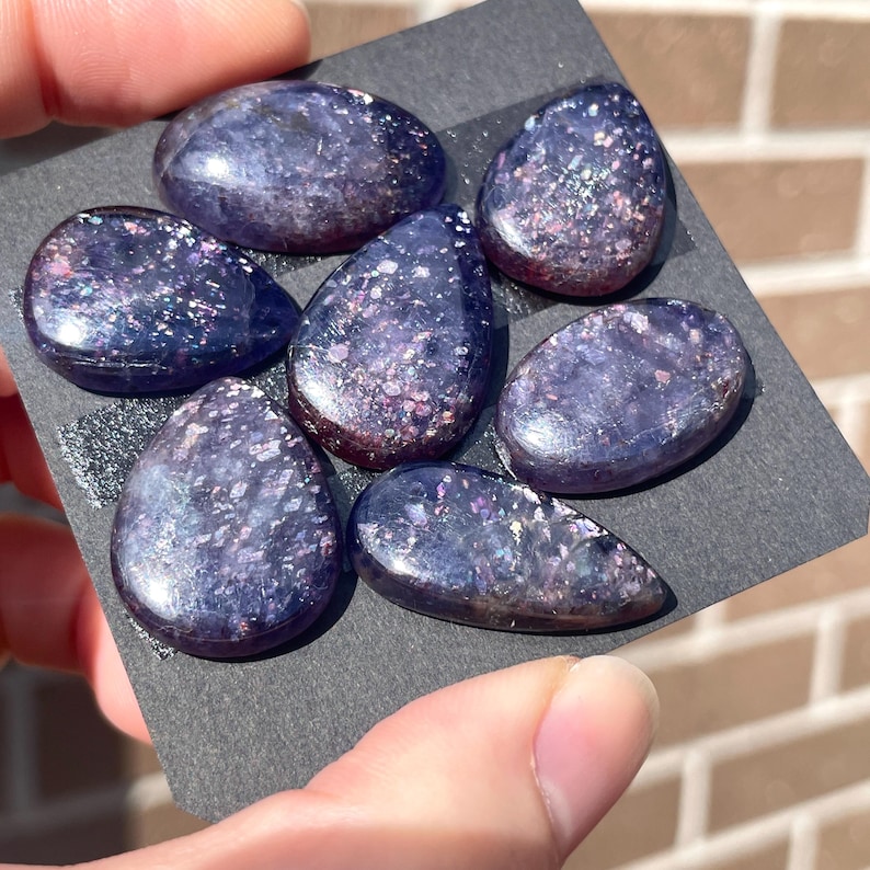 May include: Seven polished, oval-shaped stones with a deep purple hue and sparkling inclusions. The stones are arranged on a black surface, showcasing their iridescent qualities. The stones vary slightly in size and shape, offering a unique aesthetic.