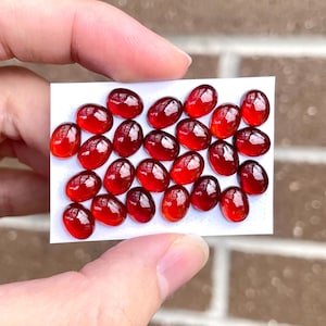 May include: A collection of 25 red oval-shaped gemstones on a white card. The gemstones are smooth and polished.