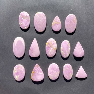 May include: A collection of 14 purple and gold cabochons, some with a teardrop shape, others oval, and some round. The cabochons are arranged in rows on a dark gray surface.