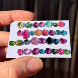 May include: A collection of small, polished, multi-colored tourmaline gemstones arranged in four horizontal rows on a white rectangular card. The stones feature various shapes, including oval, round, and teardrop, displaying shades of pink, green, blue, yellow, and black.