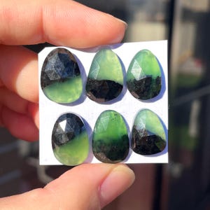 May include: Six faceted gemstones with a two-tone color scheme. The top half of each stone is a dark, opaque black, while the bottom half transitions to a vibrant green. The stones are tear-drop shaped and are displayed on a white card.