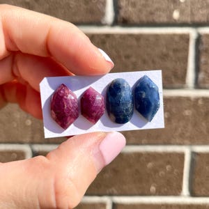 May include: Three gemstone cabochons on a white card. Two are pink and one is blue.