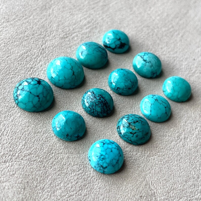 May include: Ten turquoise cabochons, each with a unique pattern of veining, are arranged in a semi-circle on a light gray surface.
