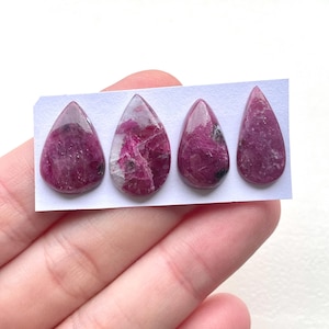May include: Four teardrop-shaped cabochons of pink ruby with white inclusions. The cabochons are arranged in a row on a white card.