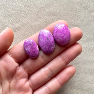 May include: Three oval-shaped purple and white cabochon gemstones. The stones are smooth and polished, with a mottled pattern of purple and white.