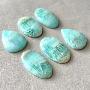 May include: A collection of six polished gemstones in shades of turquoise and pale yellow. The stones vary in shape, including ovals and a teardrop. The gemstones have a natural, swirled pattern and are displayed on a neutral surface.
