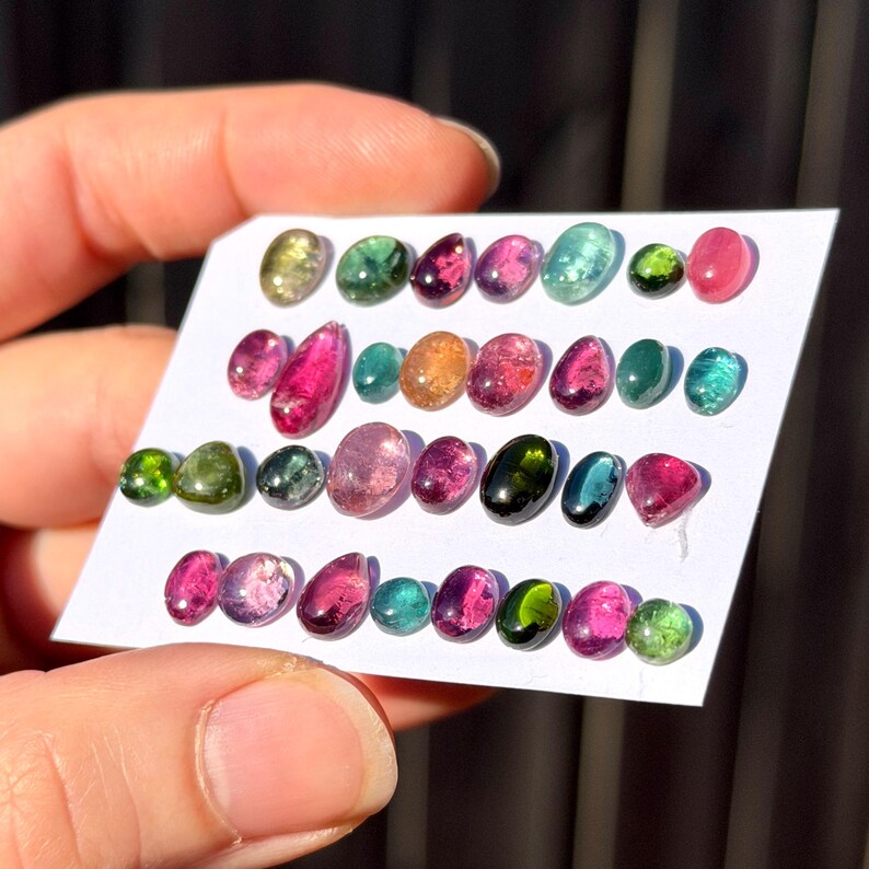 May include: A collection of small, polished, oval and teardrop-shaped tourmaline gemstones arranged in four rows on a white card. The stones display a vibrant spectrum of colors including pink, green, teal, yellow, and deep red, with translucent and opaque finishes.