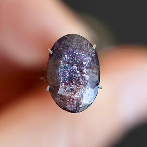 May include: An oval gemstone with a dark, speckled appearance, showcasing a shimmering, iridescent interior with purple, blue, and silver hues. The stone is secured by four silver prongs, forming a piece of jewellery.
