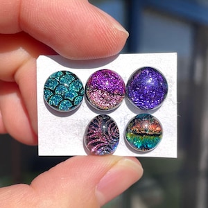 May include: Five colourful, round glass cabochons. The cabochons display diverse designs, including a teal scale pattern, sparkling purple and pink glitter, and a dark purple glitter. The bottom two cabochons have a black and pink swirl design and a colourful landscape.