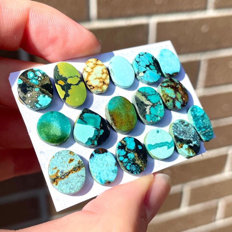 May include: A collection of turquoise gemstones in various shapes and sizes. The stones display a range of colors, including vibrant blues, greens, and browns, with unique veining patterns. The gemstones are displayed on a white surface.