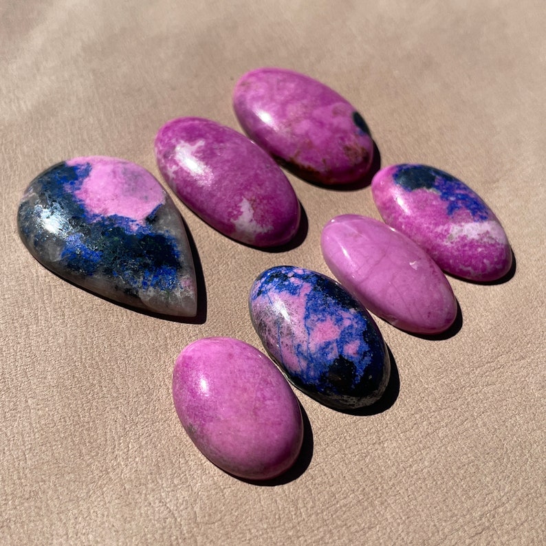 May include: Seven cabochon gemstones in various shades of pink and purple with black and blue inclusions. The stones are oval and teardrop shaped.