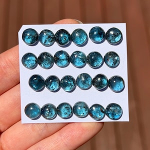 May include: A collection of twenty round, polished gemstones. Each gem is a deep teal color with dark inclusions, set on a white card. The gems are approximately 0.5 inches in diameter, and are arranged in four rows of five.