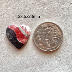 May include: A heart-shaped stone with red, black, and white patterns, measuring approximately 0.9 x 0.9 inches, next to a 20-cent coin. The stone's surface has a natural, textured appearance. The text "23.5x23mm" is visible.