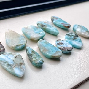 May include: A collection of ten teardrop-shaped blue and white gemstone pendants with brown and black inclusions. The stones are arranged in a row on a white surface.