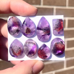 May include: A collection of eight amethyst gemstones in various shapes, including oval, teardrop, and rectangular. The gemstones display a range of purple hues with red and black inclusions, held by a hand.