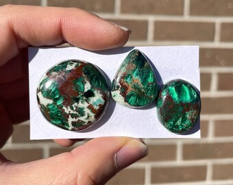 May include: Three green, white, and copper malachite cabochons. The cabochons are round, oval, and teardrop shaped.
