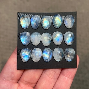 May include: A collection of 12 iridescent moonstone cabochons, each with a unique, faceted shape. The stones are arranged in two rows of six on a black background.