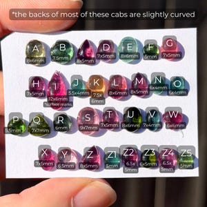 May include: A collection of loose, polished cabochon gemstones arranged on a white card, labeled A through Z5. Each stone features unique colors including pink, green, and yellow, with specific metric dimensions printed below each lettered label.