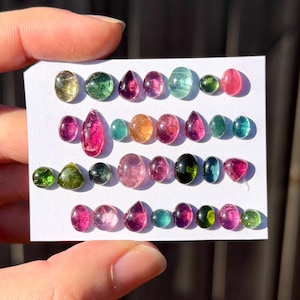May include: A collection of small, polished, multi-coloured gemstones arranged in four rows on a white card. The stones feature various shapes including ovals, rounds, and teardrops, displaying shades of pink, green, blue, yellow, and black.