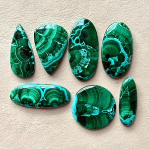Pu&ograve; includere: Una collezione di sette pietre preziose in malachite lucida di varie forme, tra cui goccia, ovale e circolare. Ogni pietra presenta tonalit&agrave; verde intenso con intricati motivi a bande vorticose e una finitura superficiale liscia.