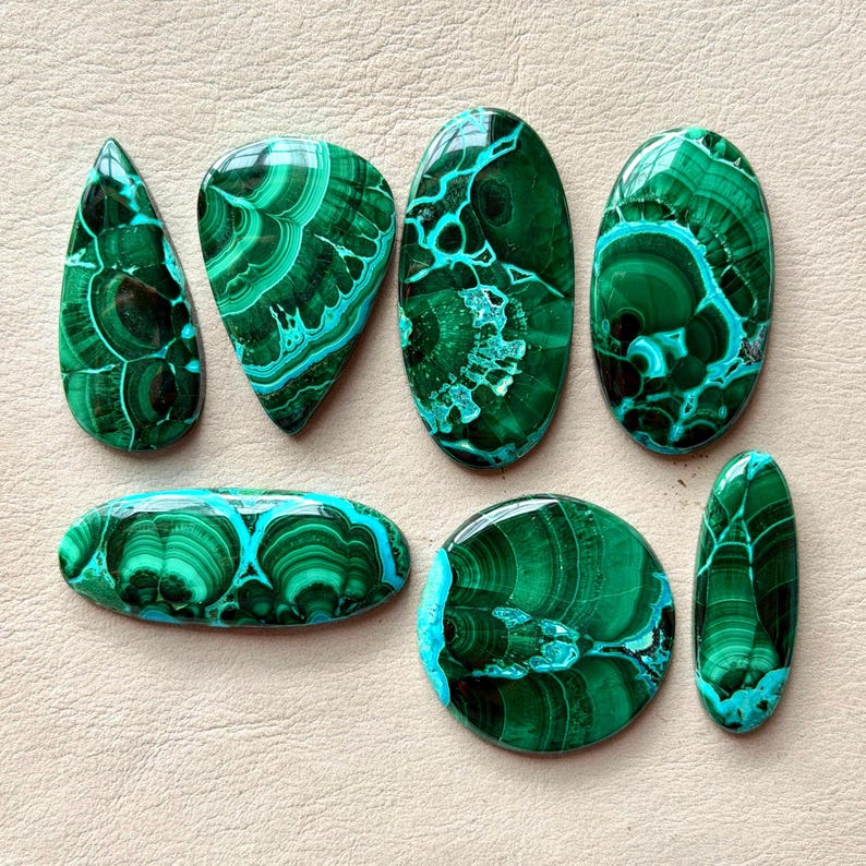 Pu&ograve; includere: Una collezione di sette pietre preziose in malachite lucida di varie forme, tra cui goccia, ovale e circolare. Ogni pietra presenta tonalit&agrave; verde intenso con intricati motivi a bande vorticose e una finitura superficiale liscia.