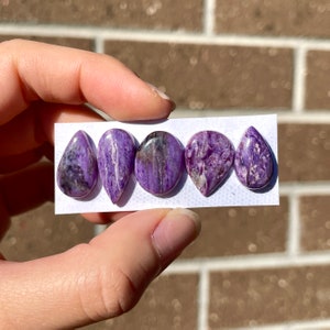 May include: Five purple and white cabochon stones in various shapes, including teardrop and oval, arranged on a white card.