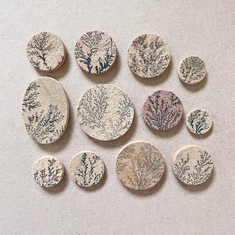 Op de afbeelding: Een verzameling van 12 ronde en ovale onderzetters met een beige achtergrond en een zwart dendritisch patroon. De onderzetters zijn gemaakt van natuursteen en hebben een rustieke, aardse uitstraling.