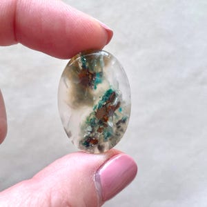 May include: A translucent oval cabochon with a white, brown, and green agate pattern. The agate has a unique, swirling pattern with hints of copper.