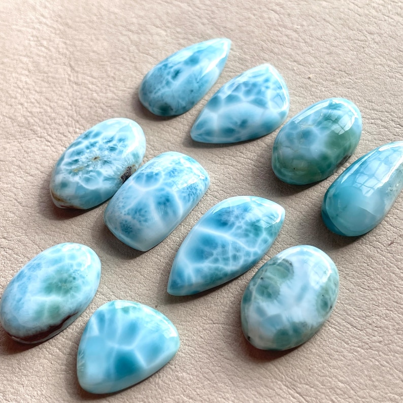 May include: A collection of ten blue and white cabochon gemstones with a marbled pattern. The stones are various shapes and sizes, and are arranged on a light brown surface.
