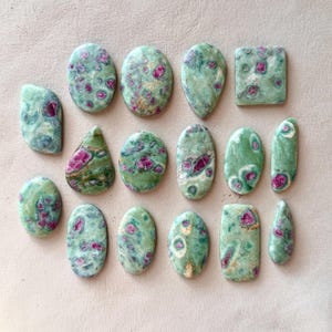 May include: A collection of polished ruby in zoisite gemstones in various shapes, including oval, teardrop, and rectangular. The stones feature a mottled green background with vibrant pink and red ruby inclusions. The gemstones are displayed on a light beige surface.