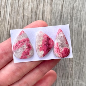 May include: Three pink and white gemstone cabochons in a teardrop shape. The cabochons are set on a white card.