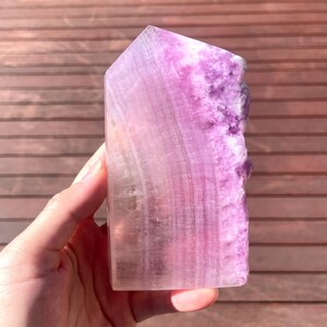 May include: A large, natural, purple and white fluorite crystal point. The crystal is polished and has a smooth, glassy surface. The crystal is a natural formation and has unique striations and banding.