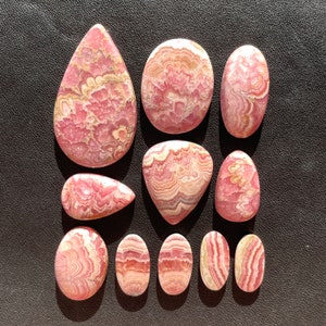 May include: A collection of 11 pink and white cabochon gemstones with a swirling pattern. The stones are various shapes and sizes, including oval, teardrop, and heart.