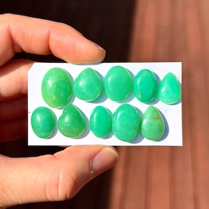 May include: A collection of ten loose, polished, bright green chrysoprase gemstones arranged in two rows on a white rectangular card. The stones feature a mix of smooth oval and teardrop shapes with a glossy, translucent finish and vibrant color.