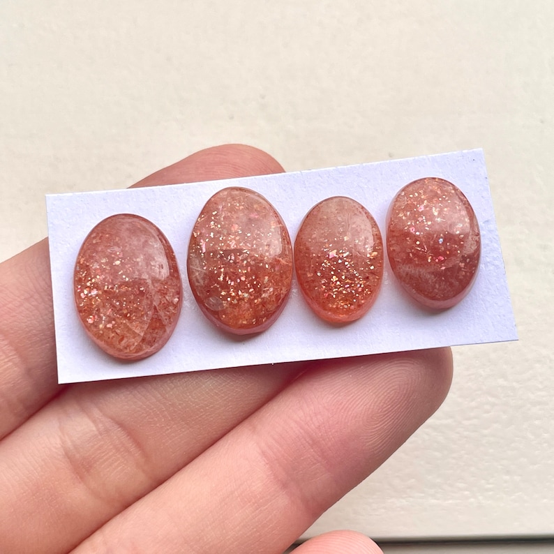 Peut inclure: Quatre cabochons ovales en pierre de soleil avec une finition chatoyante et iris&eacute;e. Les pierres sont de couleur orange clair avec des reflets dor&eacute;s et roses.