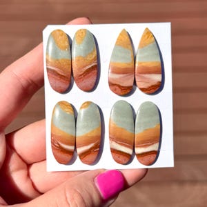 May include: Eight polished gemstone cabochons in various shapes. The stones display a layered pattern of orange, red, white, and grey-green hues. The cabochons are presented on a white card, highlighting their natural colours and textures.
