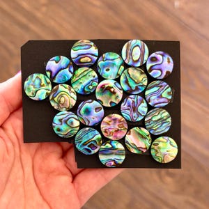 May include: A collection of iridescent, round abalone shell cabochons in shades of blue, green, purple, and gold. The cabochons are arranged on a black card, showcasing their natural patterns and colors. These decorative pieces are ideal for jewelry making or craft projects.