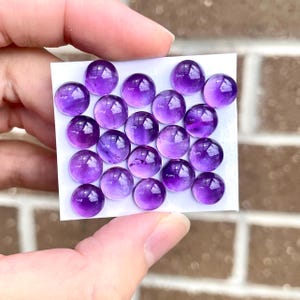 May include: A collection of 20 round, purple amethyst cabochons, arranged in a grid pattern on a white card.